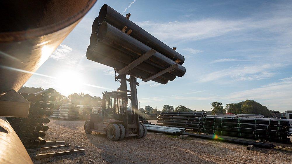 Heavy machinery carries bundle of pipes with bright sun in the background.